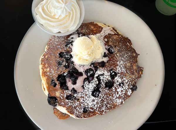 Three blueberry pancakes from Kula Bistro in Kula, Hawaii. It was topped with powdered sugar, fresh blueberries, and blueberry syrup from the island. On the side is a bowl of homemade whipped cream. This dish costs $13.15. 