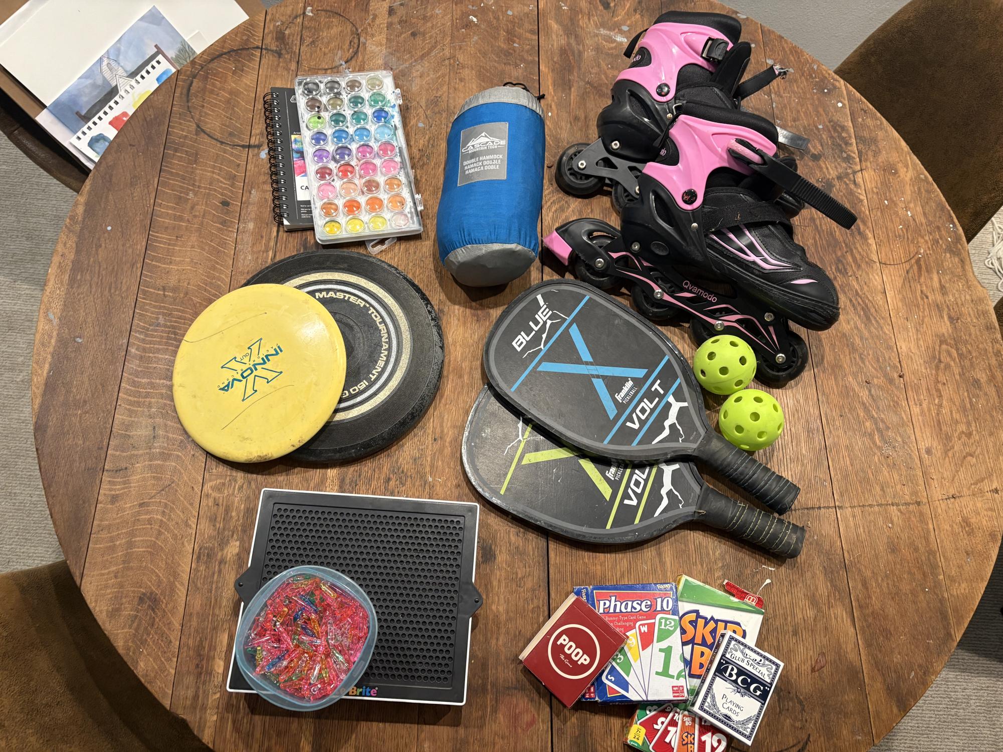 From left to right, top to bottom, a watercolor palette, hammock, roller skates,  frisbee and pickle ball set sit spread on a table. In five minutes of walking around my house, I was able to find these 6 free activities, and even a few not pictured. 